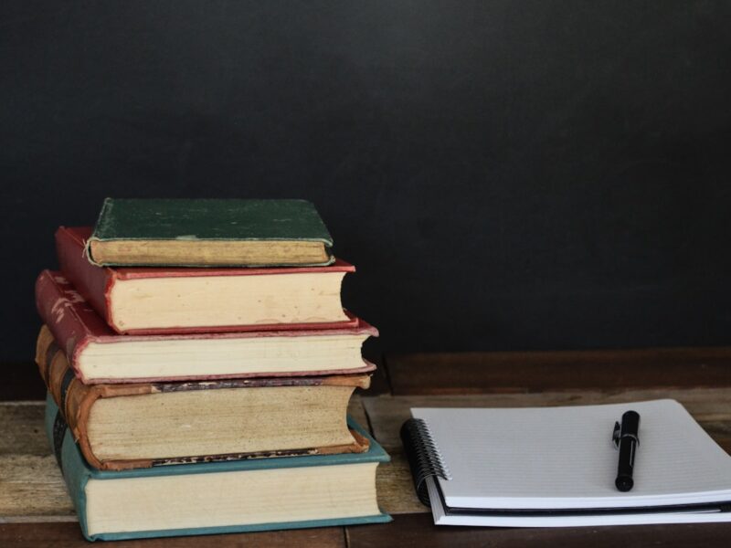 A stack of books beside a printed manuscript marked with handwritten notes on a wooden desk under warm lamplight