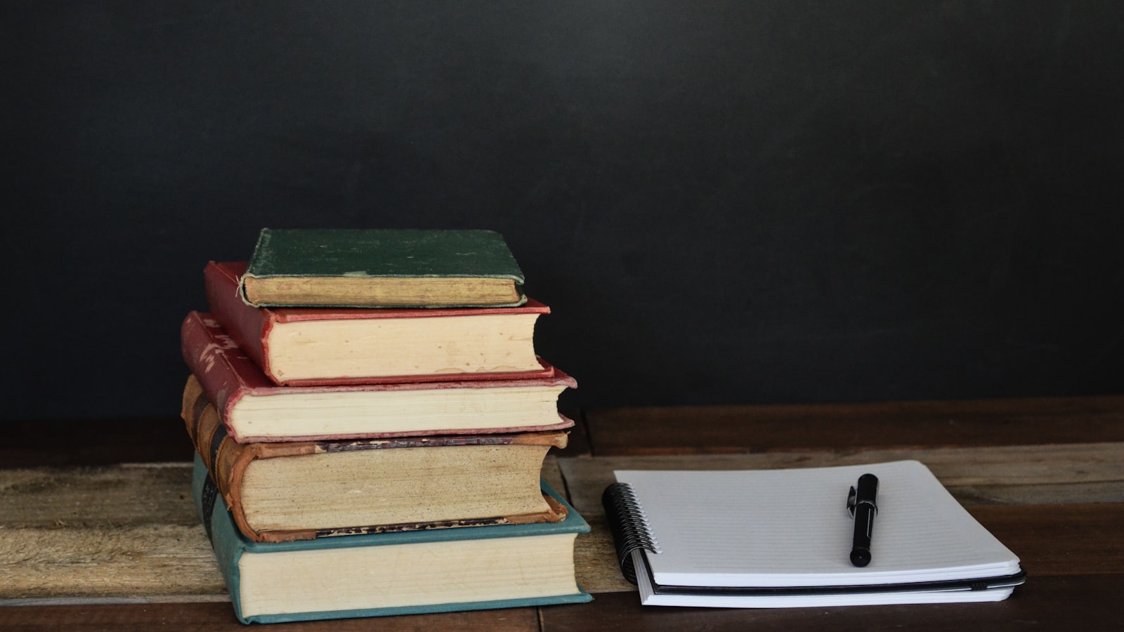 A stack of books beside a printed manuscript marked with handwritten notes on a wooden desk under warm lamplight