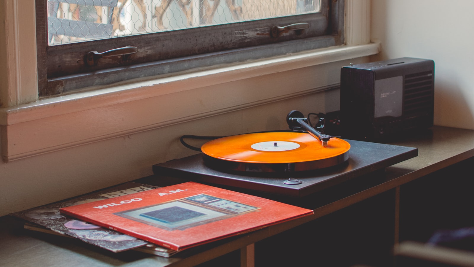 A record player on a shelf with a plain vinyl album sleeve leaning beside it in warm indoor light