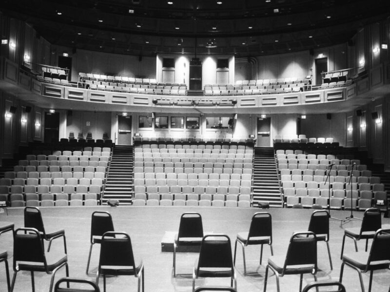 An empty concert hall stage arranged with orchestral chairs and music stands under warm lighting, with rows of empty seats beyond