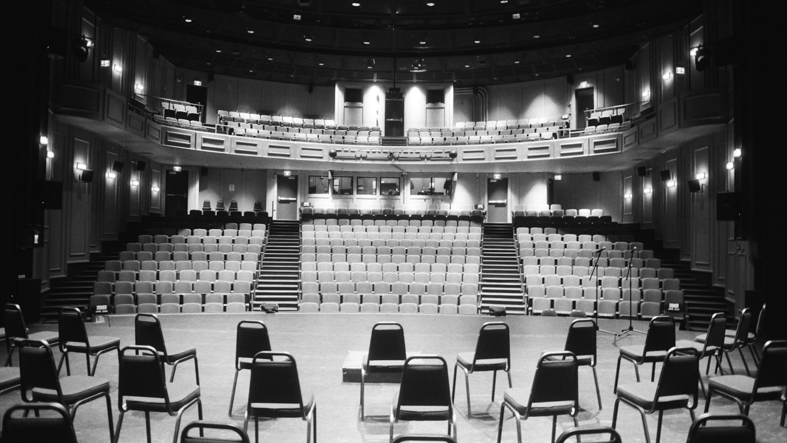 An empty concert hall stage arranged with orchestral chairs and music stands under warm lighting, with rows of empty seats beyond