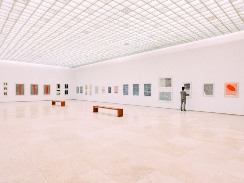 A quiet museum gallery with high ceilings, framed works along the walls and a single empty bench beneath soft overhead light