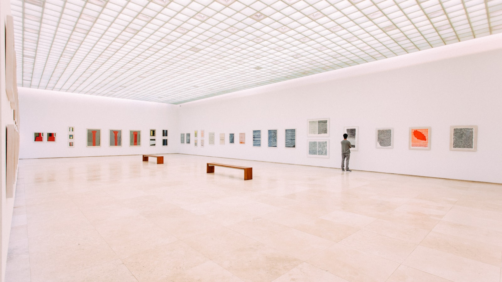 A quiet museum gallery with high ceilings, framed works along the walls and a single empty bench beneath soft overhead light