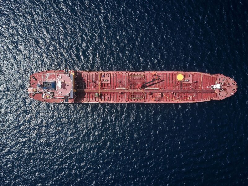AI-generated illustration of an oil tanker navigating a narrow maritime strait at dusk, with a dramatic red sky reflecting on dark water suggesting geopolitical tension