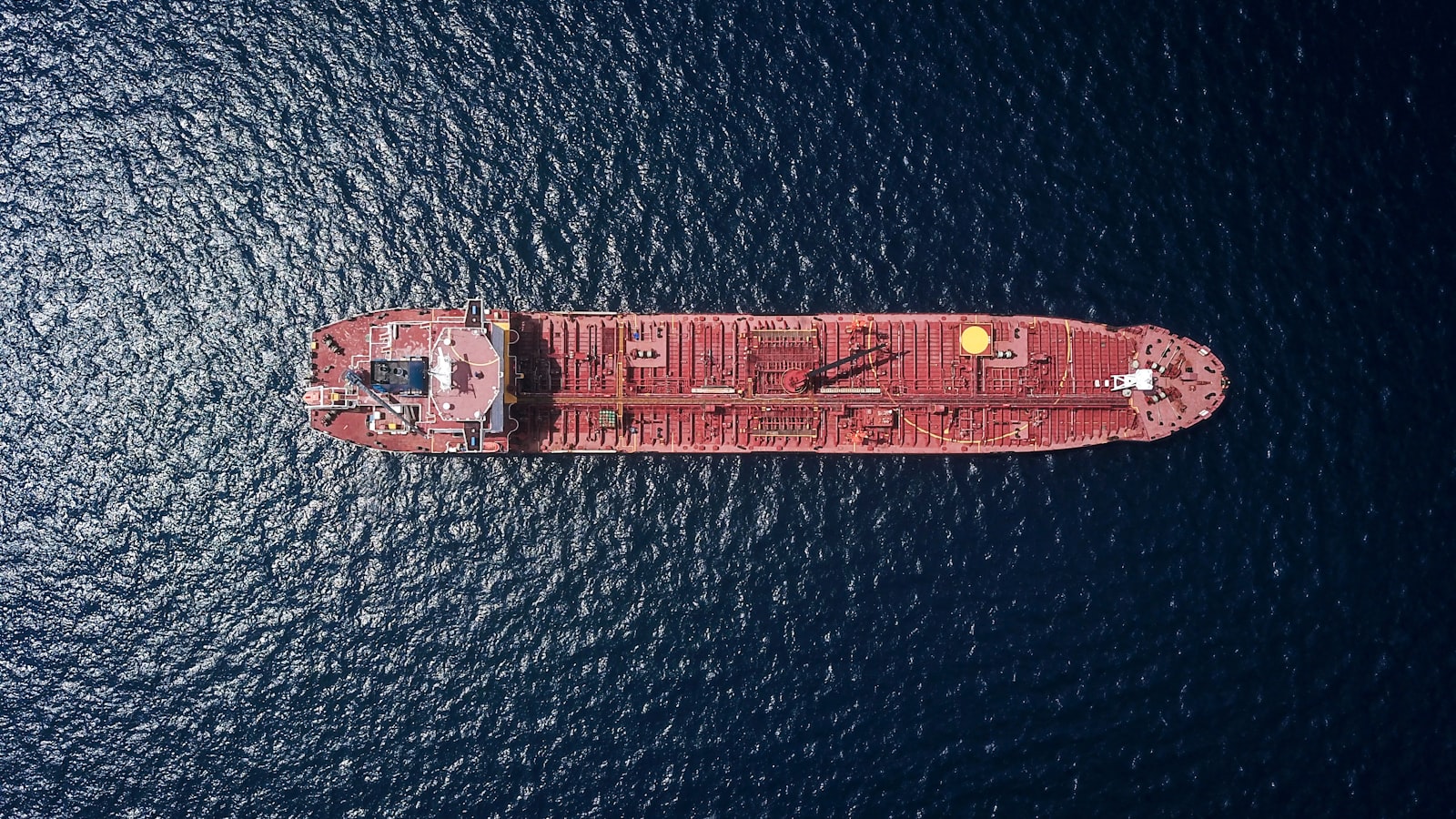AI-generated illustration of an oil tanker navigating a narrow maritime strait at dusk, with a dramatic red sky reflecting on dark water suggesting geopolitical tension