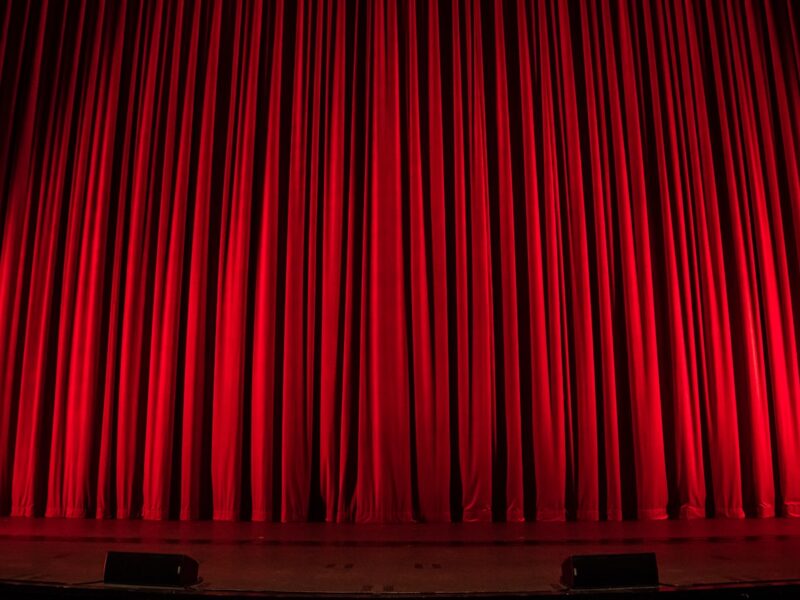 An empty theatre stage seen from the auditorium, with warm spotlights falling on a bare floor between heavy curtains