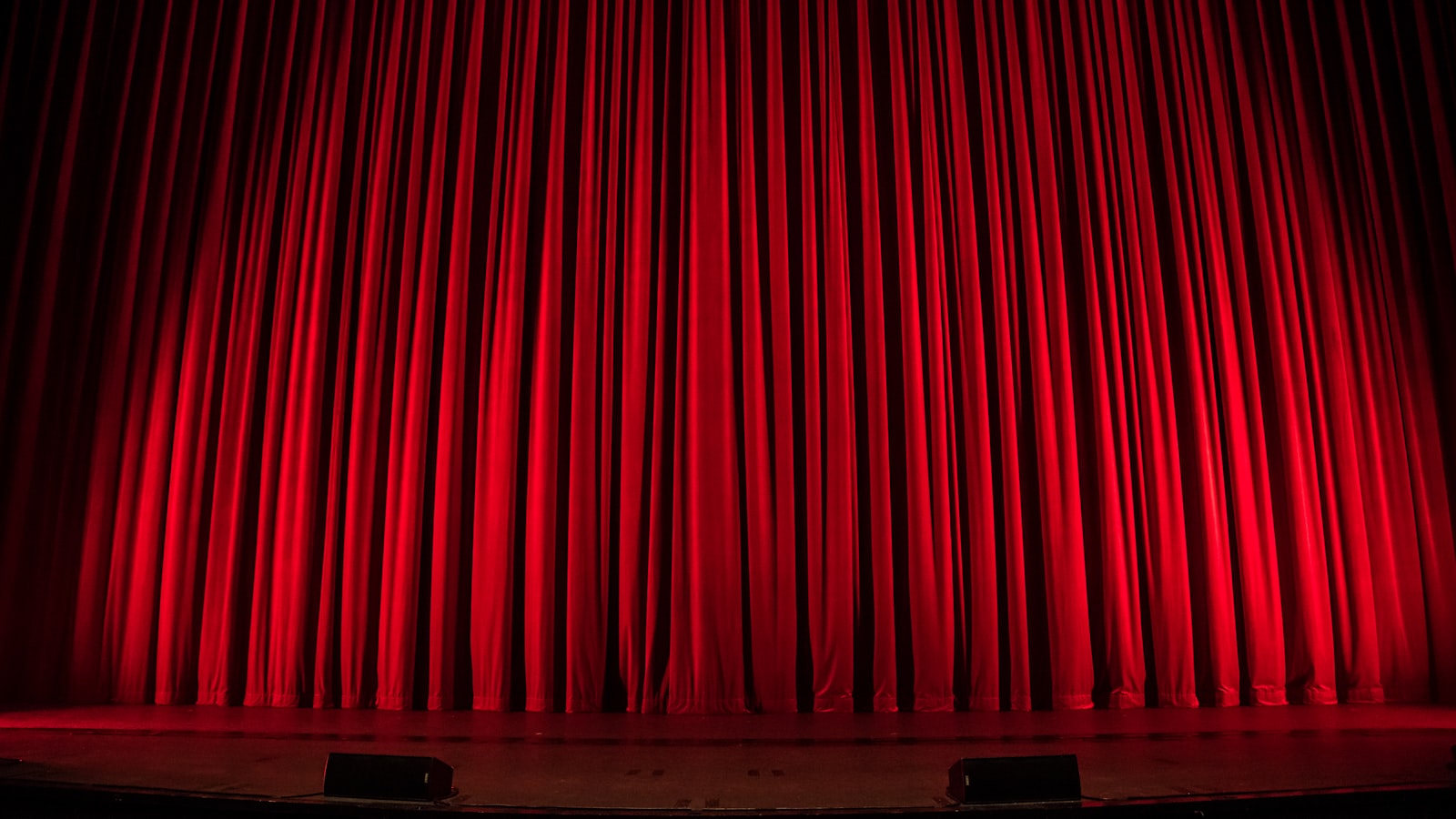 An empty theatre stage seen from the auditorium, with warm spotlights falling on a bare floor between heavy curtains