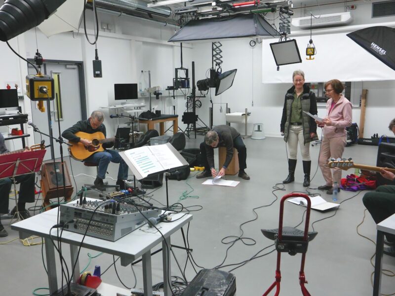 An empty rehearsal room with a wooden floor, scattered chairs and a table holding scripts, lit by daylight through tall windows