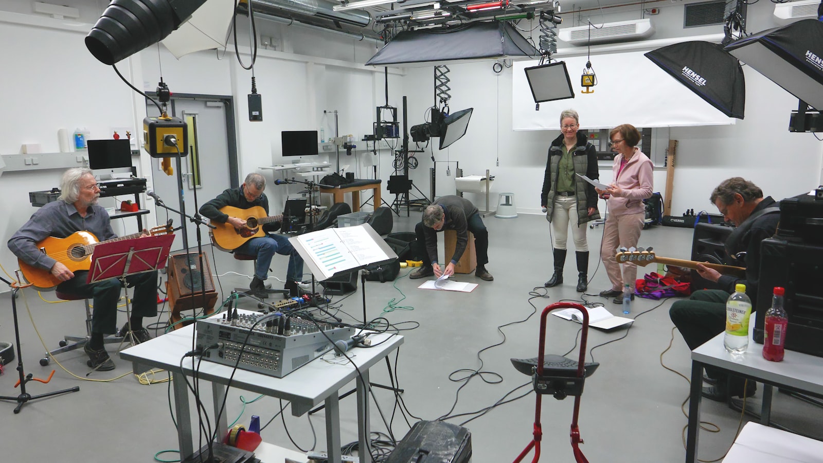 An empty rehearsal room with a wooden floor, scattered chairs and a table holding scripts, lit by daylight through tall windows